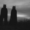 Two silhouetted figures in costumes stand in a field during twilight, creating a mysterious scene.