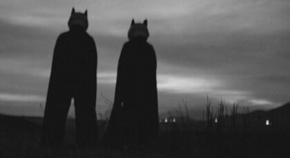 Two silhouetted figures in costumes stand in a field during twilight, creating a mysterious scene.