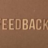 A close-up of a wooden 'FEEDBACK' sign on a textured cardboard background.
