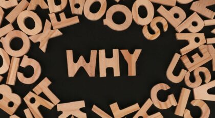 Scattered wooden alphabet letters with the word WHY at the center on a black background in a flat lay arrangement.