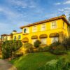 Sunny view of a Mediterranean-style villa surrounded by lush greenery and blue sky.