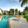 A serene poolside view with sunloungers and villas surrounded by palm trees and lush greenery.