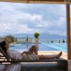 A woman in sunglasses relaxes on a sunlounger by a luxury poolside with a scenic view.